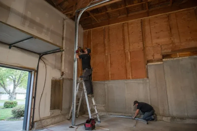 One person on a stepladder working beside a tall curved garage-door track while another kneels beside loose track sections in an unfinished garage. — High-Lift Garage Door Conversion, Innisfil